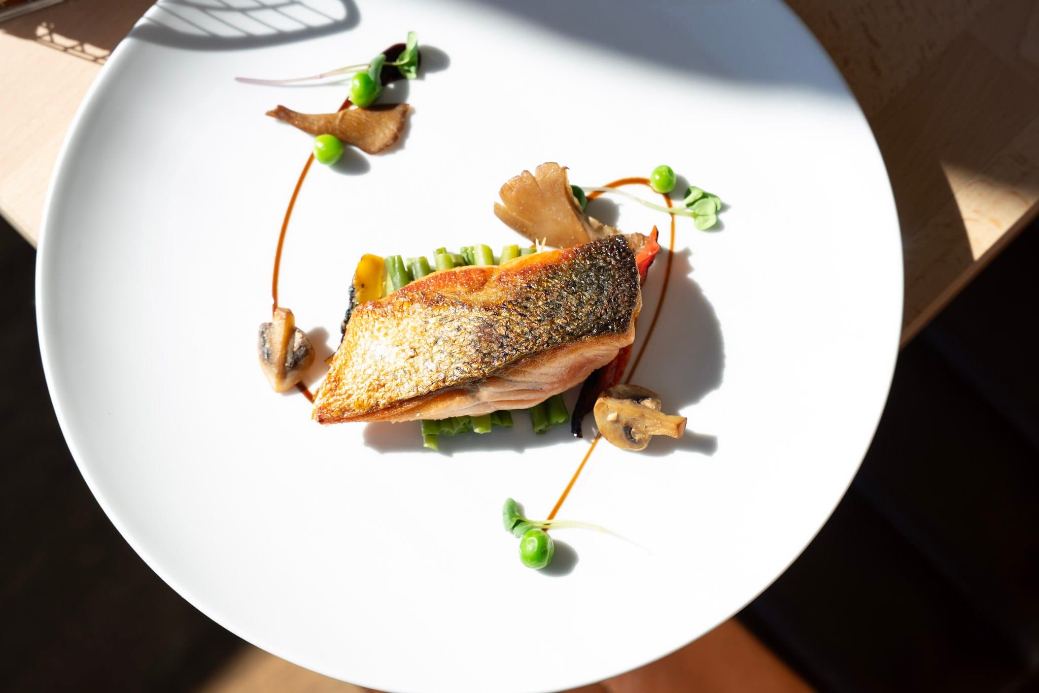 Filet de poisson à la peau croustillante sur des haricots verts, avec champignons, petits pois et un trait de sauce, dans une assiette blanche au soleil.